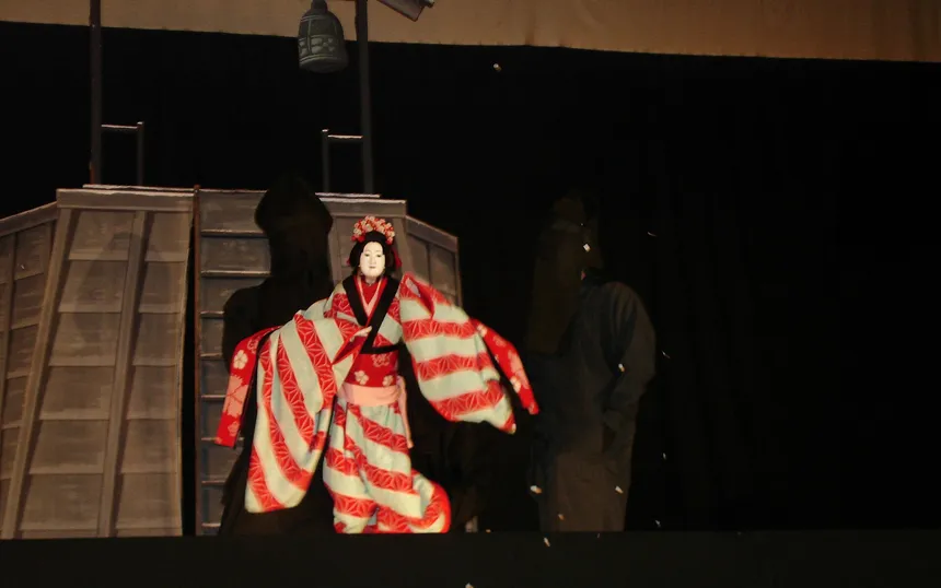 A bunraku puppet in a striped red kimono on stage, with its hooded puppeteers behind