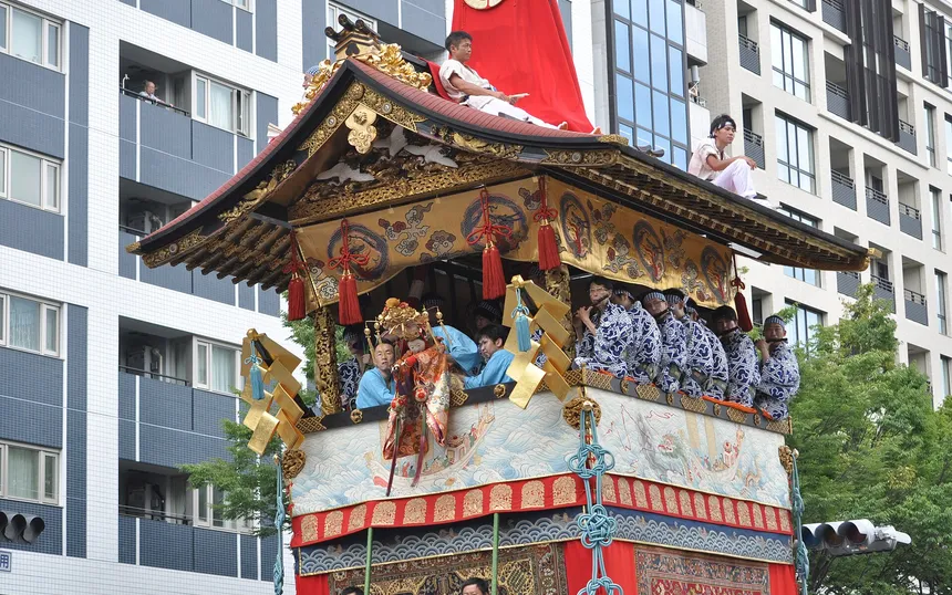 The Hoka-boko float passing between city buildings in the procession of 17 July, with men riding its roof