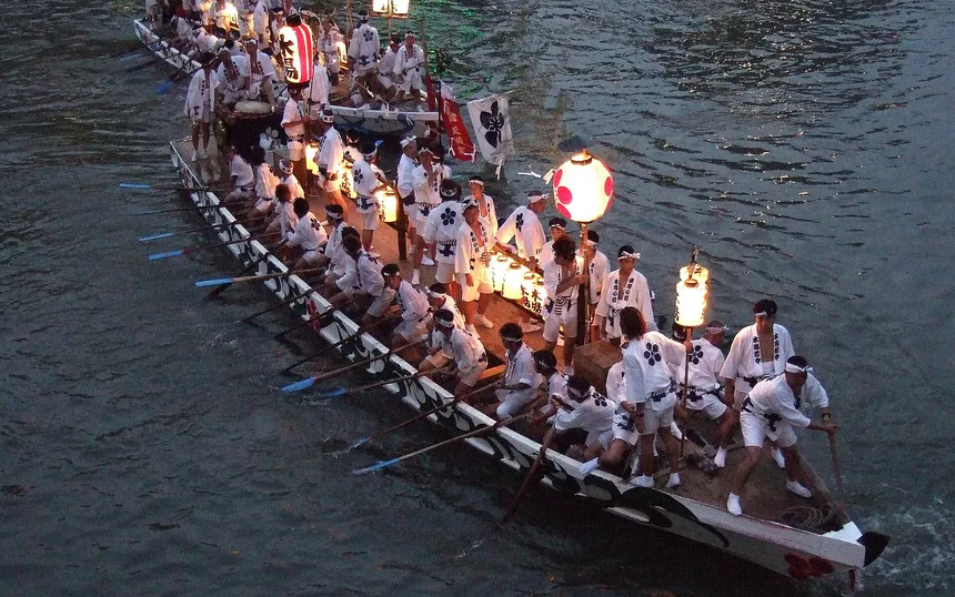 A long rowing boat of men in white coats, lit by plum-crest lanterns, on the Okawa river at dusk during the Tenjin Matsuri boat procession
