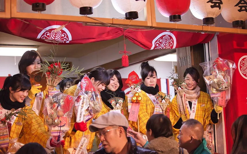 Fuku-musume in yellow kimono handing decorated lucky charms to visitors at Imamiya Ebisu Shrine