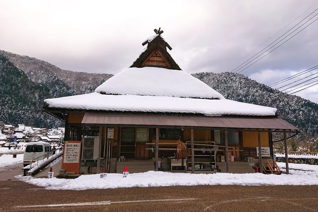 A snow-covered thatched roof at the entrance to the hamlet, with the village and hills behind